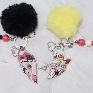 May include: Two keychains with cartoon character designs. One has a black fluffy pom-pom and the other a yellow pom-pom. Each keychain has a beaded strand with red, black, and yellow beads. The keychains have silver metal hardware.