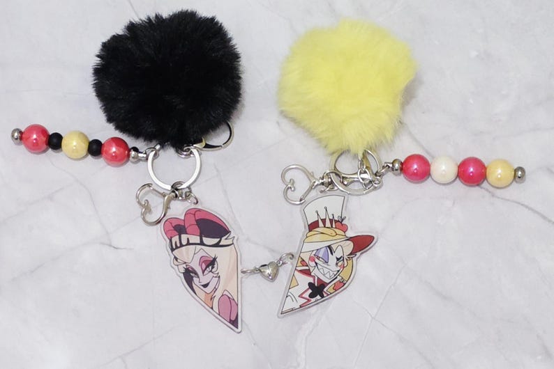 May include: Two keychains with cartoon character designs. One has a black fluffy pom-pom and the other a yellow pom-pom. Each keychain has a beaded strand with red, black, and yellow beads. The keychains have silver metal hardware.