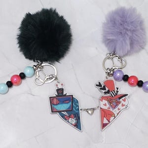 May include: Two keyrings with cartoon character designs. One has a black fluffy pom-pom, the other a purple pom-pom. Each keyring has a beaded strand with pink, blue, and black beads, and a silver-coloured keyring.