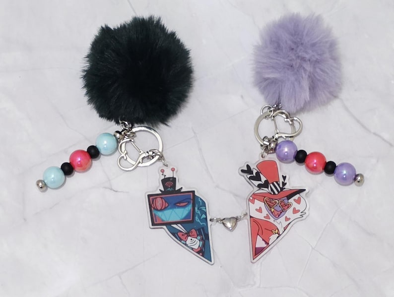 May include: Two keyrings with cartoon character designs. One has a black fluffy pom-pom, the other a purple pom-pom. Each keyring has a beaded strand with pink, blue, and black beads, and a silver-coloured keyring.