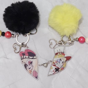 May include: Two keychains featuring cartoon character designs. One has a black fluffy pom-pom, the other a yellow one. Each keychain has a beaded strand with red, yellow, and white beads. The keychains have silver-coloured hardware.