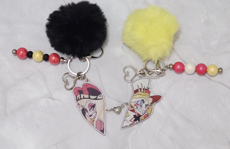 May include: Two keychains featuring cartoon character designs. One has a black fluffy pom-pom, the other a yellow one. Each keychain has a beaded strand with red, yellow, and white beads. The keychains have silver-coloured hardware.