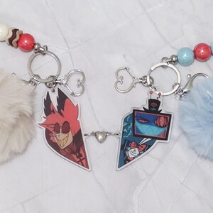 May include: Two keyrings linked by a small heart charm. Each keyring features a cartoon character charm, a fluffy pom-pom, and a beaded accent. One pom-pom is cream-coloured, the other is light blue. The beads are red, white, and blue.