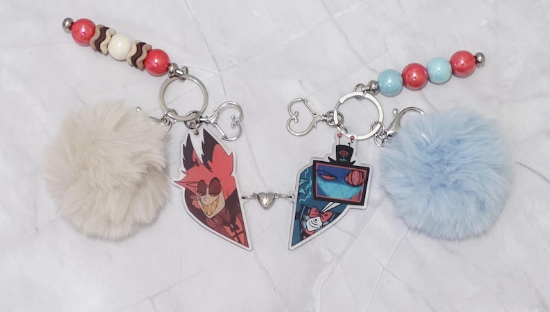 May include: Two keyrings linked by a small heart charm. Each keyring features a cartoon character charm, a fluffy pom-pom, and a beaded accent. One pom-pom is cream-coloured, the other is light blue. The beads are red, white, and blue.