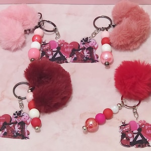 May include: Keychains with fluffy pom-poms in shades of pink and red. Each keychain features a beaded strand with white, pink, and red beads, and a charm with a graphic design and the text "F**k Me".