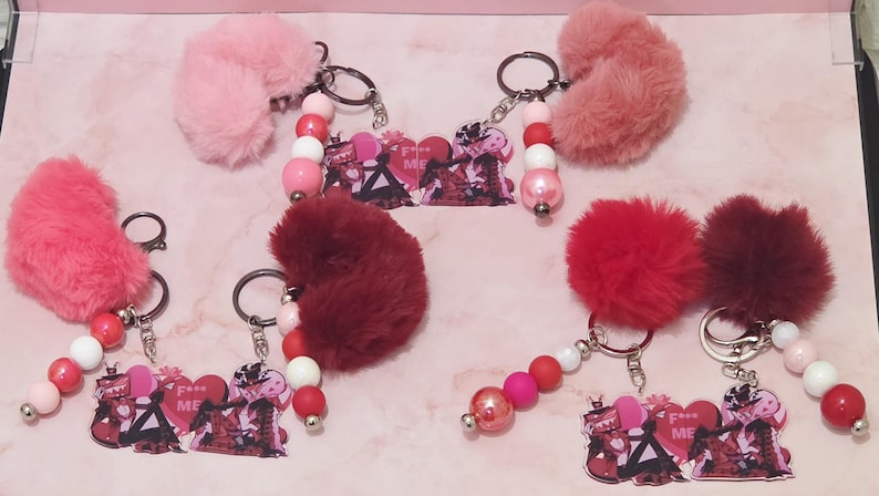 May include: Keychains with fluffy pom-poms in shades of pink and red. Each keychain features a beaded strand with white, pink, and red beads, and a charm with a graphic design and the text "F**k Me".