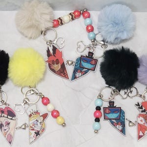 May include: A collection of keyrings with cartoon character designs, each featuring a fluffy pom-pom in colours like black, yellow, and blue. Each keyring has a metal ring and a beaded accent. The cartoon characters are in a heart shape.