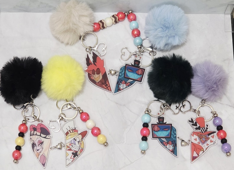 May include: A collection of keyrings with cartoon character designs, each featuring a fluffy pom-pom in colours like black, yellow, and blue. Each keyring has a metal ring and a beaded accent. The cartoon characters are in a heart shape.