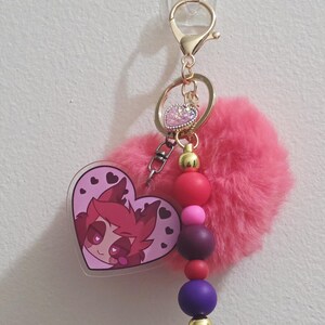 May include: A pink keychain with a heart-shaped charm featuring a cartoon character. The keychain includes a fluffy pink pom-pom, gold-colored hardware, and a beaded accent with red, pink, purple, and gold beads. The charm has a clear border.