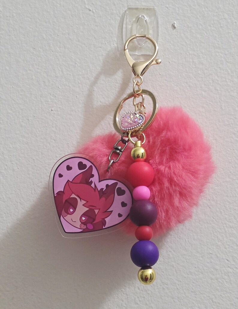 May include: A pink keychain with a heart-shaped charm featuring a cartoon character. The keychain includes a fluffy pink pom-pom, gold-colored hardware, and a beaded accent with red, pink, purple, and gold beads. The charm has a clear border.