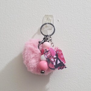 May include: A pink fluffy keychain with a metal ring, pink and white beads, and a charm featuring a cartoon character with the letters "F" and "M". The keychain is hanging on a white wall.