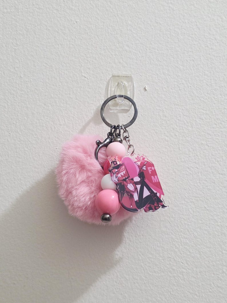 May include: A pink fluffy keychain with a metal ring, pink and white beads, and a charm featuring a cartoon character with the letters "F" and "M". The keychain is hanging on a white wall.
