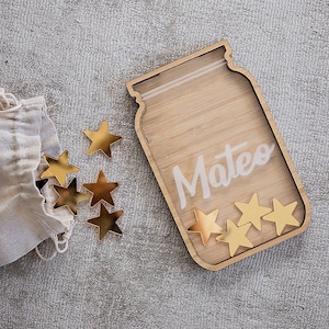 以下が含まれることがあります： 白いスクリプトで「Mateo」という名前が入った、木製の瓶型の報酬チャート。 金色の星型のトークンが小さなキャンバスバッグの周りに散らばっています。 瓶には星型の切り抜きが5つあります。