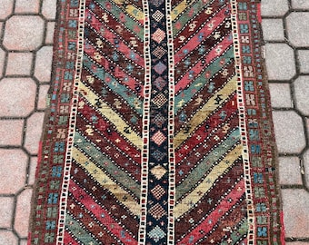 Op de afbeelding: Een vintage Turks tapijt met een geometrisch patroon in tinten rood, blauw, groen en bruin. Het tapijt heeft een franje rand en is geweven met ingewikkelde details.