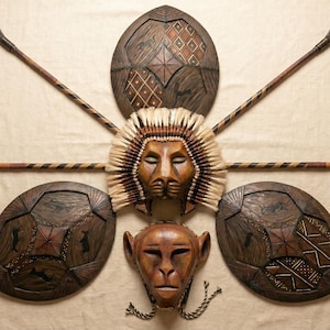 Può includere: Una collezione di arte tribale in legno fatta a mano, tra cui una maschera di leone, una maschera di scimmia, tre scudi e due lance. I pezzi marroni presentano intagli e motivi intricati, disposti su uno sfondo beige.
