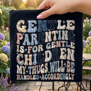 Puede incluir: Un cojín cuadrado negro con el texto "GENTLE PARENTING IS FOR GENTLE CHILDREN MY THUGS WILL BE HANDLED ACCORDINGLY" en varias fuentes estampadas. El cojín está sostenido por una mano en un jardín.