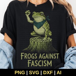 May include: Black t-shirt featuring a green frog wearing a bandana, sitting on a tree stump, with the text "Frogs Against Fascism" in a distressed font. The frog is raising a fist. The bottom of the image shows "PNG | SVG | DXF | AI".