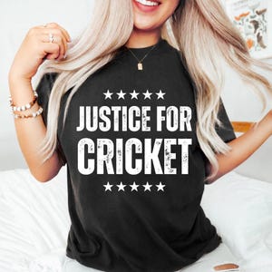 May include: Black t-shirt with the text "JUSTICE FOR CRICKET" in white, with five stars above and below the text. The shirt is a crew neck and appears to be made of a soft material.