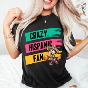 May include: Black t-shirt with the words "CRAZY. HISPANIC. FAN" in green, pink, and yellow brushstrokes. A cartoon fox wearing a sombrero and holding a basketball is featured below the text.