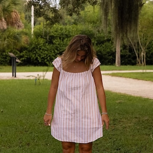 May include: A short-sleeved dress featuring a white base with vertical orange stripes, a square neckline, and a relaxed, straight-cut silhouette that falls above the knees. The garment has gathered cap sleeves and a simple, unadorned hemline.