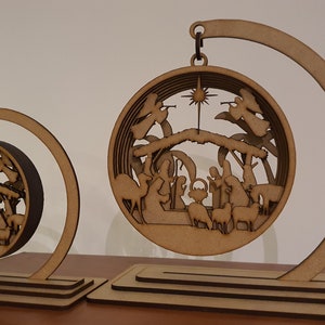 May include: Two wooden Christmas ornaments depicting the Nativity scene. The ornaments are round and feature a detailed cutout of the manger, Mary, Joseph, baby Jesus, angels, and animals. The ornaments are suspended from a wooden stand.