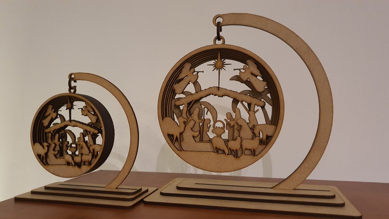 May include: Two wooden Christmas ornaments depicting the Nativity scene. The ornaments are round and feature a detailed cutout of the manger, Mary, Joseph, baby Jesus, angels, and animals. The ornaments are suspended from a wooden stand.