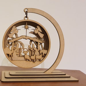May include: A wooden Christmas ornament depicting the nativity scene with angels, a manger, and animals. The ornament is suspended from a wooden stand.