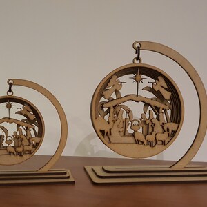 May include: Two wooden Christmas ornaments depicting the nativity scene. The ornaments are circular and feature a layered design with a star at the top. The ornaments are suspended from a curved wooden stand.