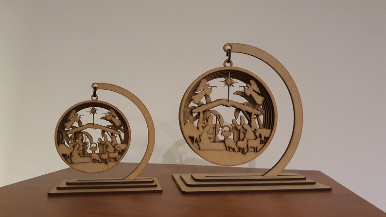 May include: Two wooden Christmas ornaments depicting the nativity scene. The ornaments are circular and feature a layered design with a star at the top. The ornaments are suspended from a curved wooden stand.