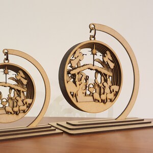 May include: Two wooden Christmas ornaments depicting the Nativity scene. The ornaments are suspended from a crescent-shaped stand. The larger ornament is in the foreground and the smaller ornament is in the background.