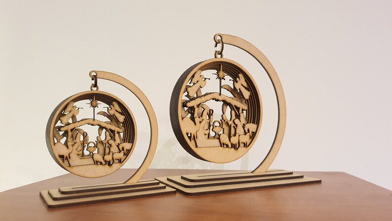 May include: Two wooden Christmas ornaments depicting the Nativity scene. The ornaments are suspended from a crescent-shaped stand. The larger ornament is in the foreground and the smaller ornament is in the background.