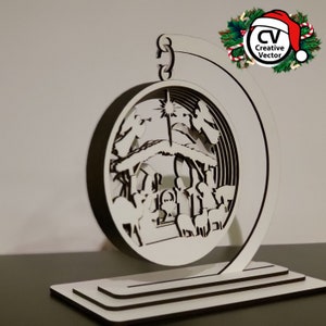 May include: White wooden Christmas ornament featuring a detailed laser-cut nativity scene with a star, angels, and animals. The ornament is suspended from a curved stand.