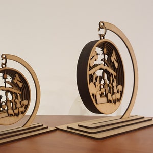 May include: Two wooden Christmas ornaments depicting the nativity scene. The ornaments are circular and feature a detailed cutout design. They are displayed on wooden stands.