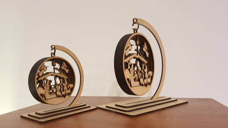 May include: Two wooden Christmas ornaments depicting the nativity scene. The ornaments are circular and feature a detailed cutout design. They are displayed on wooden stands.