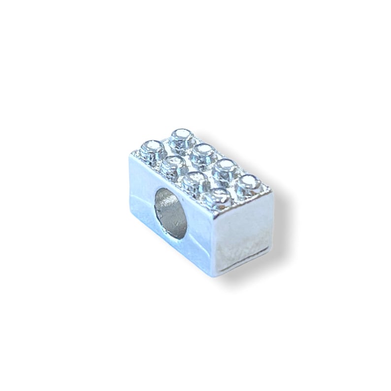 May include: A silver charm in the shape of a Lego brick with a hole in the center. The brick is decorated with small, clear rhinestones.