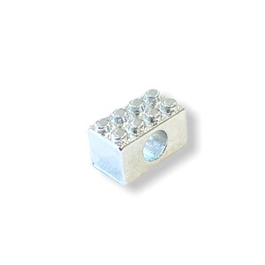 May include: Silver charm in the shape of a Lego brick. The charm has a hole in the center for attaching to a bracelet or necklace.