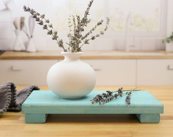May include: A white ceramic vase with dried lavender flowers sits on a teal blue wooden pedestal with a rustic finish. The pedestal has two legs and is made of wood.