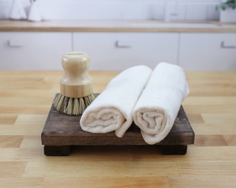 May include: Two white hand towels rolled up and placed on a dark brown wooden tray with a wooden brush with natural bristles.