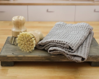 May include: Two wooden handled brushes with natural bristles, one with a brown bristle and one with a tan bristle, are on a wooden tray with a gray waffle weave towel.