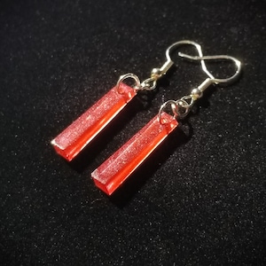 Puede incluir: Un par de pendientes de gota rojos con purpurina y ganchos plateados.