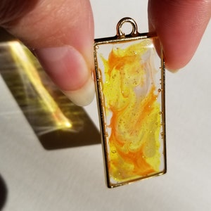 May include: A gold-framed rectangular pendant with a clear resin background filled with orange and white swirls and glitter.