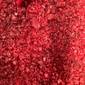 May include: Close-up of red, sparkly bath salts. The salts are small and have a rough texture.