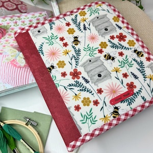 May include: A white fabric binder with a red gingham border and a red spine. The fabric features a floral pattern with bees and beehives. The binder has a red label that says "Hand Made".