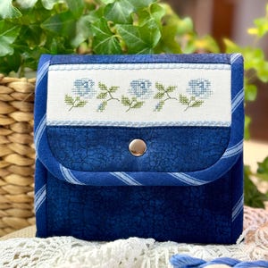 Puede incluir: Un protector de agujas de tela azul con cierre de botón blanco y un diseño bordado blanco de flores azules. El protector de agujas está sobre una superficie de crochet blanco.