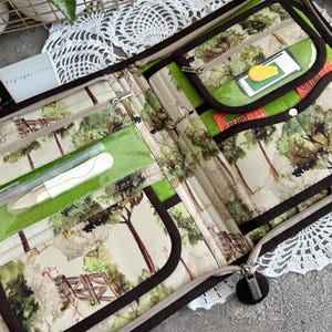 May include: A brown zippered pouch with a green and beige fabric interior. The pouch has multiple pockets and a clear plastic window. The fabric features a watercolor design of trees and a fence.