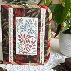 May include: A fabric-covered notebook or planner case featuring a dark brown background with a printed autumn leaf pattern. The center panel displays a cross-stitched floral design in blue and rust tones, framed by two vertical strips of white lace.