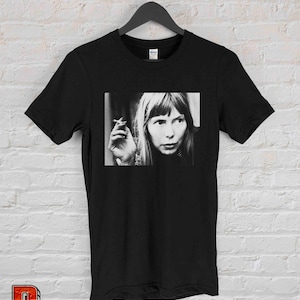 May include: Black t-shirt featuring a black and white image of a woman holding a cigarette. The image is a close-up of the woman's face and hand. The t-shirt is hanging on a black hanger against a white brick wall.