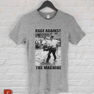 Op de afbeelding: Een grijs t-shirt met een zwart-wit afbeelding van een protestsc&egrave;ne en de tekst "RAGE AGAINST THE MACHINE" in vette, zwarte hoofdletters. De afbeelding toont politieagenten die een persoon arresteren.