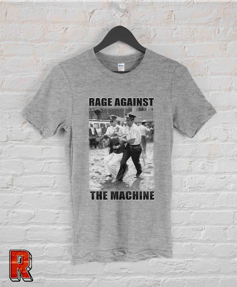 Op de afbeelding: Een grijs t-shirt met een zwart-wit afbeelding van een protestsc&egrave;ne en de tekst "RAGE AGAINST THE MACHINE" in vette, zwarte hoofdletters. De afbeelding toont politieagenten die een persoon arresteren.