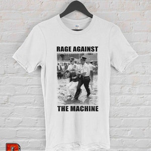 Op de afbeelding: Wit T-shirt met een zwart-wit afbeelding van een protestsc&egrave;ne en de tekst "RAGE AGAINST THE MACHINE". De afbeelding toont een persoon die door politieagenten wordt ge&euml;scorteerd. Het T-shirt hangt aan een zwarte hanger.